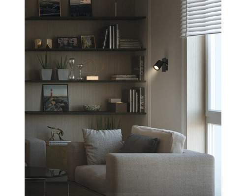 Modernes Wohnzimmer mit Stoffsofa und Wandregalen. Ein schwarzer verstellbarer LED-Wandstrahler erzeugt einen Lichtpunkt im dunklen Raum.