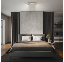 Modernes Schlafzimmer mit grauem Polsterbett, zweiflammigem Deckenstrahler, Wandpaneel in Betonoptik, Fischgrätparkett und dunklen Vorhängen.