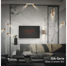 Helles, modernes Wohnzimmer mit Briloner Logo auf TV, IDA-Serie Leuchten an Marmorwand, Decke und als Stehlampe neben grauem Sofa.