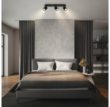 Modernes Schlafzimmer mit einer dreiflammigen Deckenleuchte, die ein graues Bett, eine Wand in Betonoptik und dunkle Vorhänge beleuchtet.