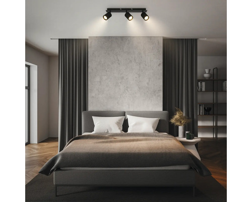 Modernes Schlafzimmer mit einer dreiflammigen Deckenleuchte, die ein graues Bett, eine Wand in Betonoptik und dunkle Vorhänge beleuchtet.
