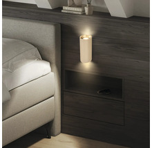 Moderne, geriffelte Wandleuchte in Zylinderform an einer dunklen Holzwand neben einem Bett, die Licht nach oben und unten strahlt.