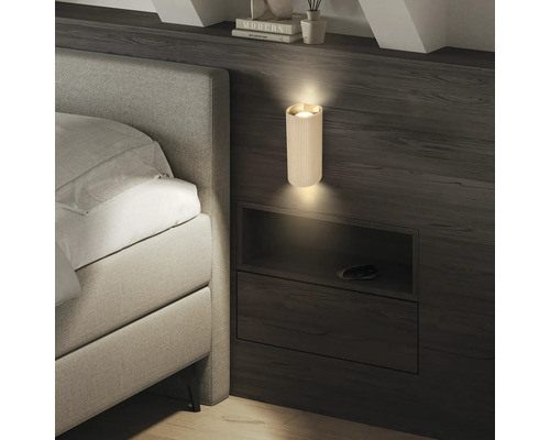 Moderne, geriffelte Wandleuchte in Zylinderform an einer dunklen Holzwand neben einem Bett, die Licht nach oben und unten strahlt.