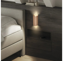 Moderne zylindrische Wandleuchte mit Lamellenstruktur an einer dunklen Holzwand neben einem hellgrauen Polsterbett.
