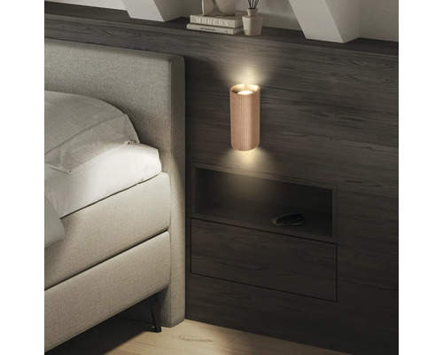 Moderne zylindrische Wandleuchte mit Lamellenstruktur an einer dunklen Holzwand neben einem hellgrauen Polsterbett.