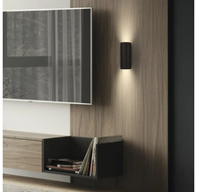 Moderne schwarze Zylinder-Wandleuchte mit Up- und Downlight auf einem Holzpaneel im Wohnzimmer neben einem Fernseher.