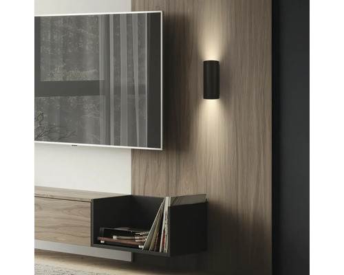 Moderne schwarze Zylinder-Wandleuchte mit Up- und Downlight auf einem Holzpaneel im Wohnzimmer neben einem Fernseher.