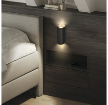 Moderne Wandleuchte mit geriffelter Textur in einem Schlafzimmer, die Licht nach oben und unten über einer Bettablage aus Holz abgibt.