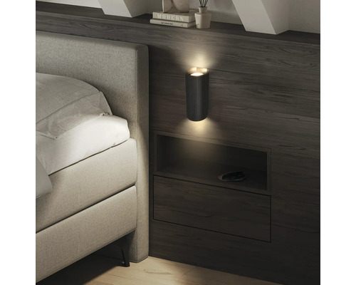 Moderne Wandleuchte mit geriffelter Textur in einem Schlafzimmer, die Licht nach oben und unten über einer Bettablage aus Holz abgibt.