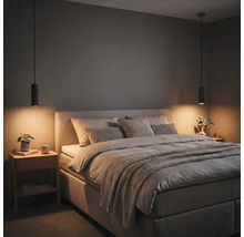 Modernes Schlafzimmer mit gepolstertem Bett, grauen Kissen, zwei schwarzen zylindrischen Pendelleuchten und einem Nachttisch aus Holz.