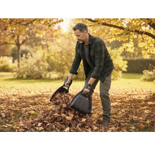 Ein Mann verwendet zwei schwarze Laubschaufeln, um trockenes Laub in einem herbstlichen Garten aufzusammeln.