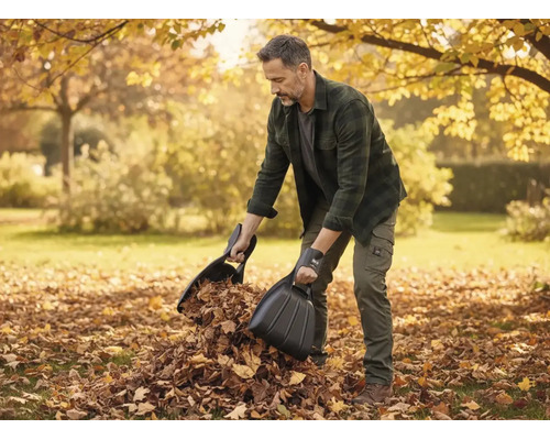 Ein Mann verwendet zwei schwarze Laubschaufeln, um trockenes Laub in einem herbstlichen Garten aufzusammeln.