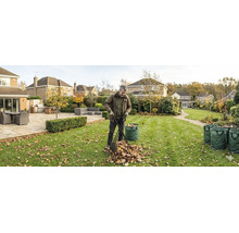 Ein Mann sammelt mit einem Laubgreifer Herbstlaub in einem Garten mit Rasen, Terrasse und mehreren grünen Gartenabfallsäcken.