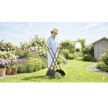Frau benutzt schwarzen Laub- und Grasschnittsammler in einem hellen Garten mit Blumenrabatten und Gartenhaus.