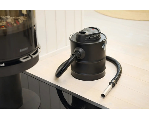 Elektrischer Aschesauger mit Metallbehälter und flexiblem Saugschlauch in einem hellen Raum neben einem Kamin.