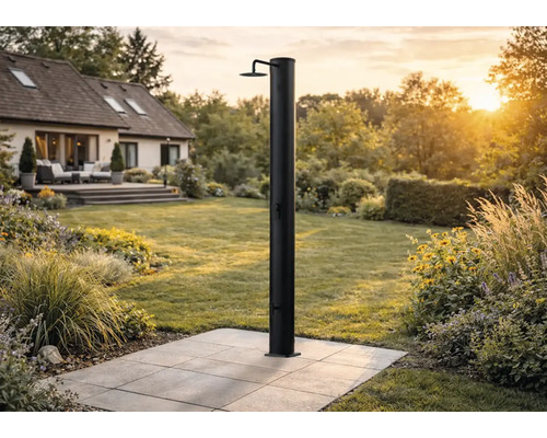 Schwarze Solardusche auf einer gepflasterten Terrasse in einem weitläufigen, hellen Garten mit Wohnhaus im Hintergrund.