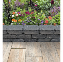 Niedrige Gartenmauer aus dunkelgrauen strukturierten Mauersteinen vor einem bunten Blumenbeet und hellen Terrassenplatten.