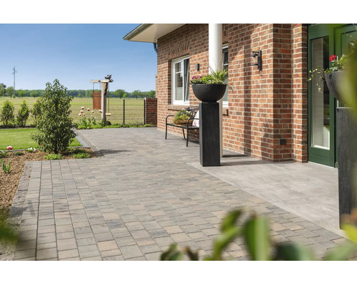 Großzügig gestaltete Terrasse mit Pflastersteinen aus Beton vor einem Backsteinhaus, inklusive Gartenbank und dekorativem Pflanzgefäß.