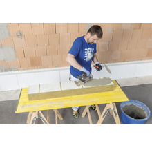 Handwerker trägt mit einer Zahnspachtel Klebemörtel auf eine Dämmplatte aus Mineralwolle auf einem Arbeitstisch auf.