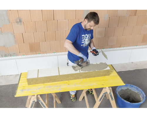 Handwerker trägt mit einer Zahnspachtel Klebemörtel auf eine Dämmplatte aus Mineralwolle auf einem Arbeitstisch auf.