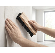 Eine Person glättet beim Tapezieren eine Tapete mit einer Rautner Tapezierbürste mit Holzgriff und dunklen Borsten an einer Wand.