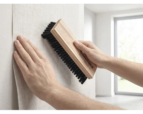 Eine Person glättet beim Tapezieren eine Tapete mit einer Rautner Tapezierbürste mit Holzgriff und dunklen Borsten an einer Wand.