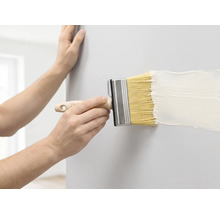 Eine Person streicht eine Wand mit einem breiten Flächenstreicher und weißer Farbe.