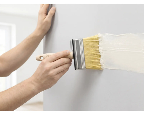 Eine Person streicht eine Wand mit einem breiten Flächenstreicher und weißer Farbe.