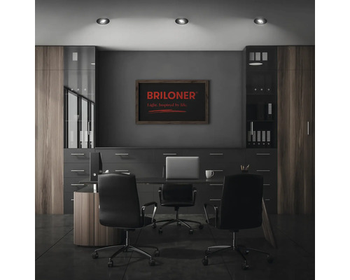 Modernes Büro mit Schreibtisch, zwei Bürostühlen, Schrankwand und drei Deckenleuchten sowie Briloner Logo an der Wand.