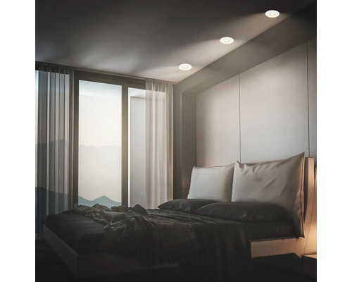 Modernes Schlafzimmer mit Einbaustrahlern in der Decke, die ein warmes Licht auf das Bett werfen, vor einer großen Fensterfront.