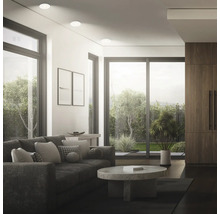 Modernes Wohnzimmer mit Einbaustrahlern, beigem Sofa, rundem Steincouchtisch und großen Fensterfronten mit Blick in den Garten.