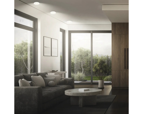 Modernes Wohnzimmer mit Einbaustrahlern, beigem Sofa, rundem Steincouchtisch und großen Fensterfronten mit Blick in den Garten.