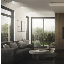 Modernes Wohnzimmer mit Einbaustrahlern in der Decke, großem Sofa, Rundtisch und Fensterfront mit Blick in den Garten.