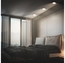 Modernes Schlafzimmer mit Einbaustrahlern in der Decke, die ein warmes Licht auf das Bett werfen, vor einer großen Fensterfront.