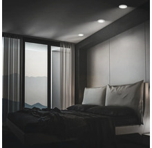 Modernes Schlafzimmer mit Einbauleuchten an der Decke, bodentiefem Fenster und minimalistischem Bett vor einer Wandverkleidung.