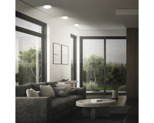 Modernes Wohnzimmer mit Einbaustrahlen an der Decke, großen Fenstern, Sofa und rundem Couchtisch mit Blick in einen Garten.