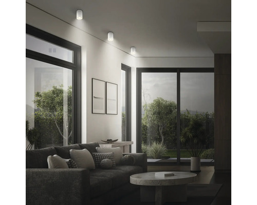 Modernes Wohnzimmer mit drei zylindrischen Deckenstrahlern an der Wand, große Fensterfronten und gemütliche Sofaecke.