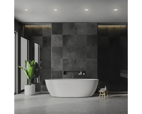Modernes Badezimmer mit freistehender Badewanne, dunklen Fliesen und indirekter Beleuchtung, ergänzt durch Zimmerpflanze und Holzschemel.