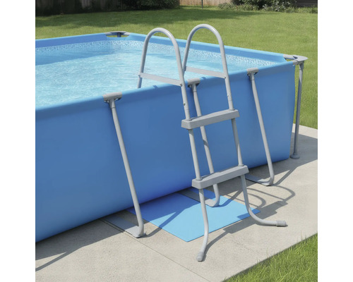 Rechteckiger Aufstellpool in Blau mit grauer Poolleiter in einem hellen Garten auf einer Betonfläche.
