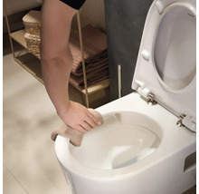 Eine Hand reinigt den Rand einer weißen wandhängenden Toilette mit einem beigen Tuch in einem hellen Badezimmer.