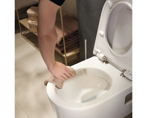 Eine Hand reinigt den Rand einer weißen wandhängenden Toilette mit einem beigen Tuch in einem hellen Badezimmer.
