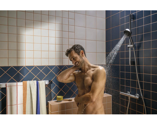 Mann beim Duschen unter einer Brausegarnitur in einem modern gefliesten Badezimmer mit Handtuchhalter und Handtüchern.