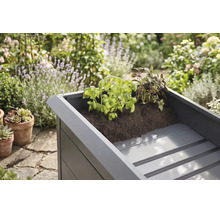 Hochbeet aus Metall mit Kräutern in Erde bepflanzt, im Hintergrund ein sonniger Garten mit verschiedenen Pflanzen.