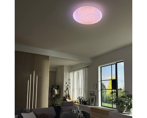 Runde LED-Deckenleuchte mit Sterneneffekt in einem hellen Wohnzimmer mit Holzlamellenwand, Sofa und großen Fenstern.