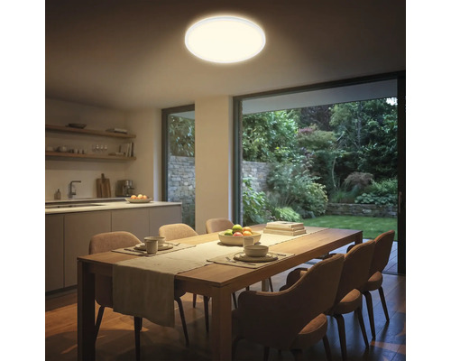 Runde LED-Deckenleuchte in einem modernen Esszimmer mit Holztisch, gepolsterten Stühlen und Blick durch eine Glasfront in den Garten.