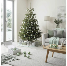 Festlich geschmückter Weihnachtsbaum in einem hellen Wohnzimmer mit Sofa, Couchtisch aus Holz sowie silbernen und grünen Christbaumkugeln.