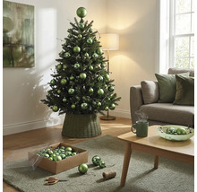 Dekorierter künstlicher Weihnachtsbaum mit grünen Kugeln in verschiedenen Größen in einem geflochtenen Ständer im hellen Wohnzimmer.