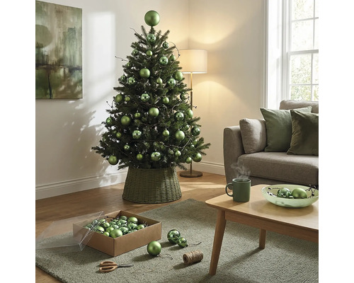 Dekorierter künstlicher Weihnachtsbaum mit grünen Kugeln in verschiedenen Größen in einem geflochtenen Ständer im hellen Wohnzimmer.