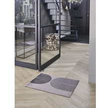 Moderne Fußmatte mit geometrischem Muster auf Fischgrät-Holzboden in hellem Flur mit Glastür und Treppe.
