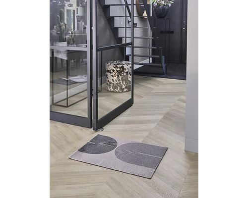 Moderne Fußmatte mit geometrischem Muster auf Fischgrät-Holzboden in hellem Flur mit Glastür und Treppe.